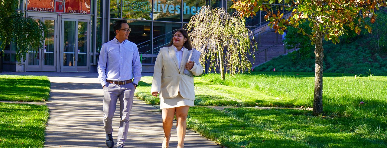 UC international students Randy Mercado and Khwahish Singh stroll in front of UC's University Pavillion building, which shows the Next Lives Here banner in its window.