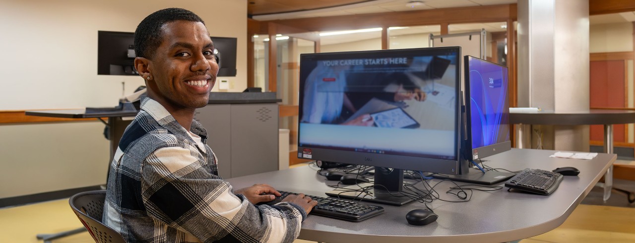 Siinboon Daba sits at a desk with a computer and smiles at the camera