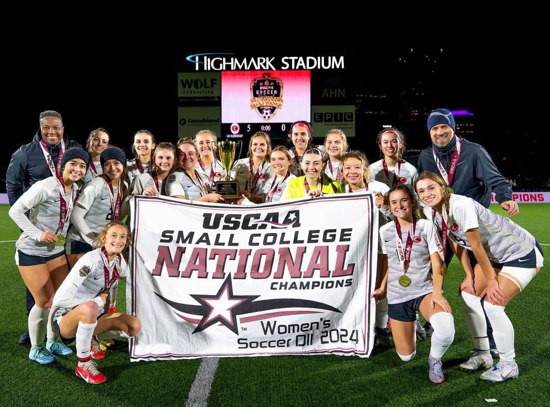 The UC Clermont women’s soccer team celebrates its 2024 national championship win by gathering around a banner celebrating the accomplishment.