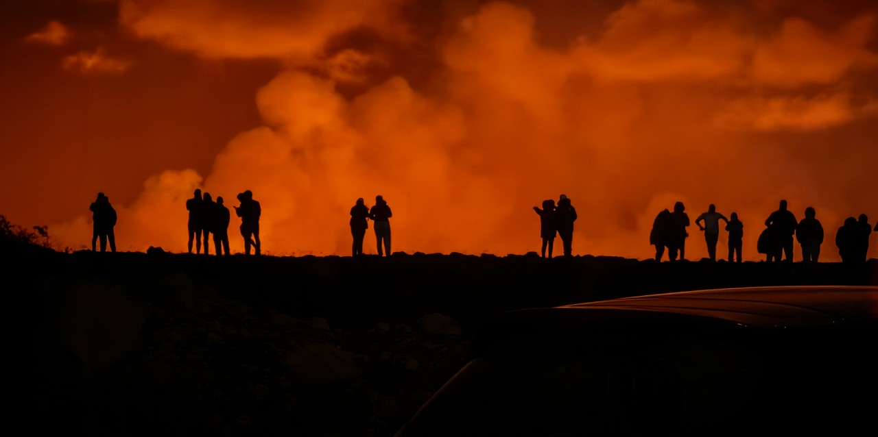 People are silhouetted against the orange glow of an eruption and cloud of gas.