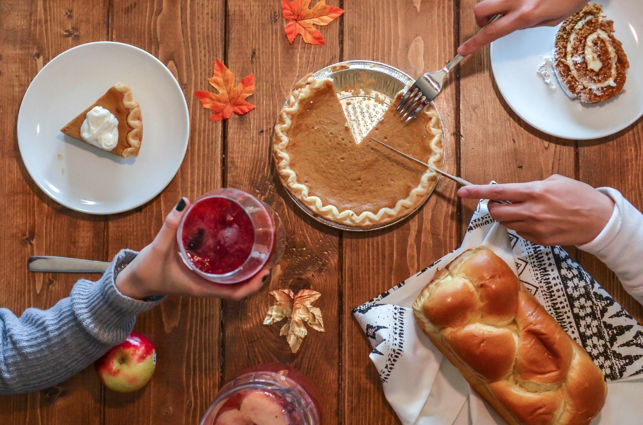 A person cuts into a pumpkin pie on a table filled with food while another person holds up a drink.