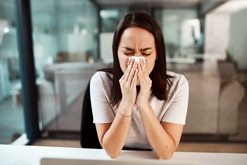 A woman blows her nose in an office setting.