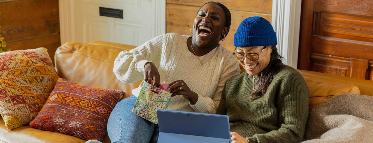 Two people, one on a laptop and one holding a gift bag, laugh while sitting on a couch.