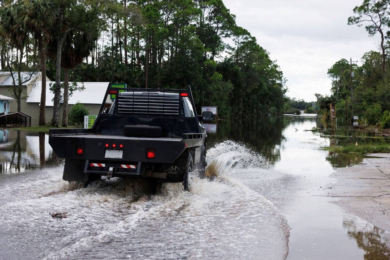 A flatbed truck drives through flooded water in a neighborhood along a river.