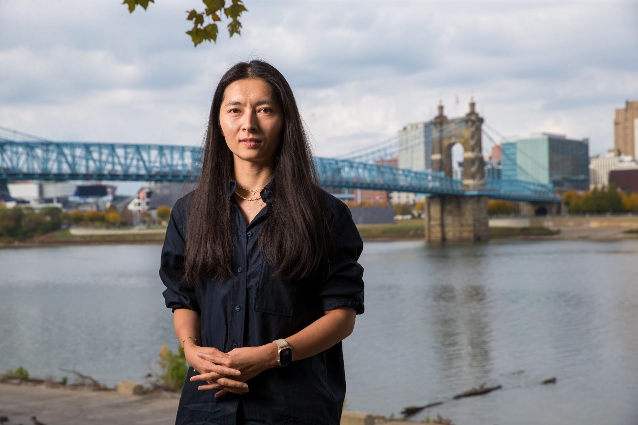 Dongmei Feng stands on the banks of the Ohio River. Behind her is the river and a bridge. 