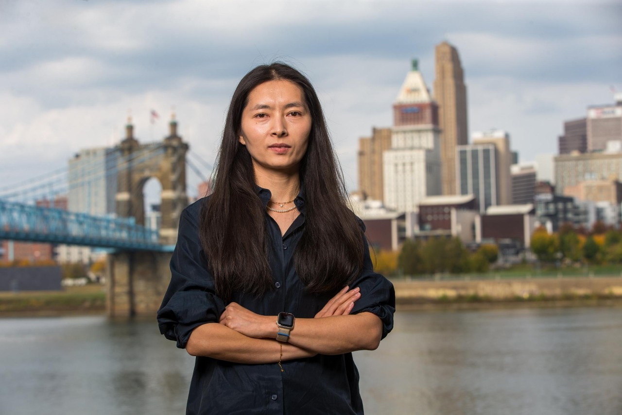 Dongmei Feng stands on the banks of the Ohio River with the Cincinnati skyline behind her.