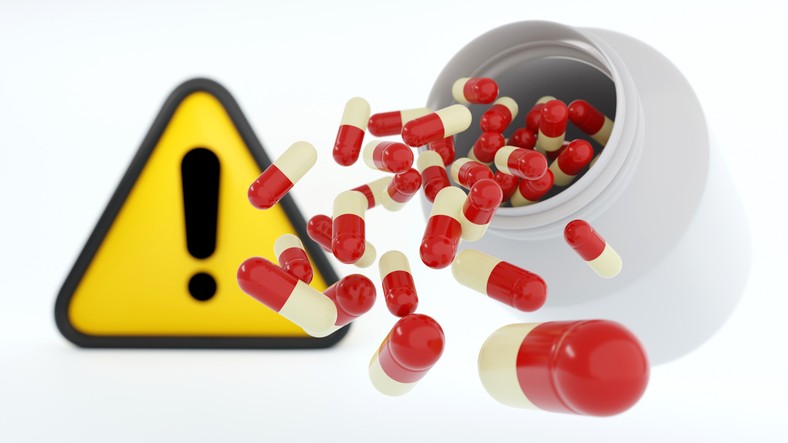 Capsule spills pouring out of a pill bottle with a yellow triangle with an exclamation point warning sign in the background